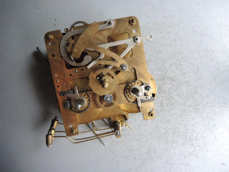May include: Close-up of a vintage clock movement. The brass-colored mechanism features numerous gears, levers, and springs, mounted on a rectangular plate. The intricate design showcases the internal workings of a clock.