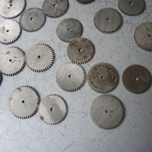 May include: A collection of small, circular metal gears with toothed edges and a square hole in the center. Some gears show signs of rust and wear, scattered on a light gray surface.