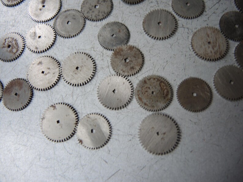 May include: A collection of small, circular metal gears with toothed edges and a square hole in the center. Some gears show signs of rust and wear, scattered on a light gray surface.