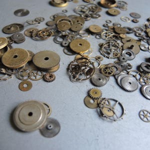 May include: A collection of vintage watch gears in various sizes and shapes, made of brass and steel. The gears are scattered across a light gray surface, showcasing their intricate designs and aged appearance. Ideal for steampunk projects or jewelry making.