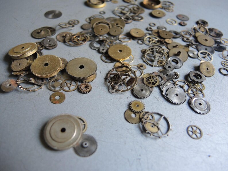 May include: A collection of vintage watch gears in various sizes and shapes, made of brass and steel. The gears are scattered across a light gray surface, showcasing their intricate designs and aged appearance. Ideal for steampunk projects or jewelry making.