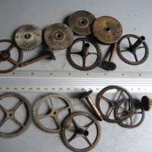 May include: A collection of vintage clock gears and components, showcasing various sizes and designs. The gears are made of metal and display a weathered, aged appearance. A ruler is placed near the gears for scale, with measurements in inches.