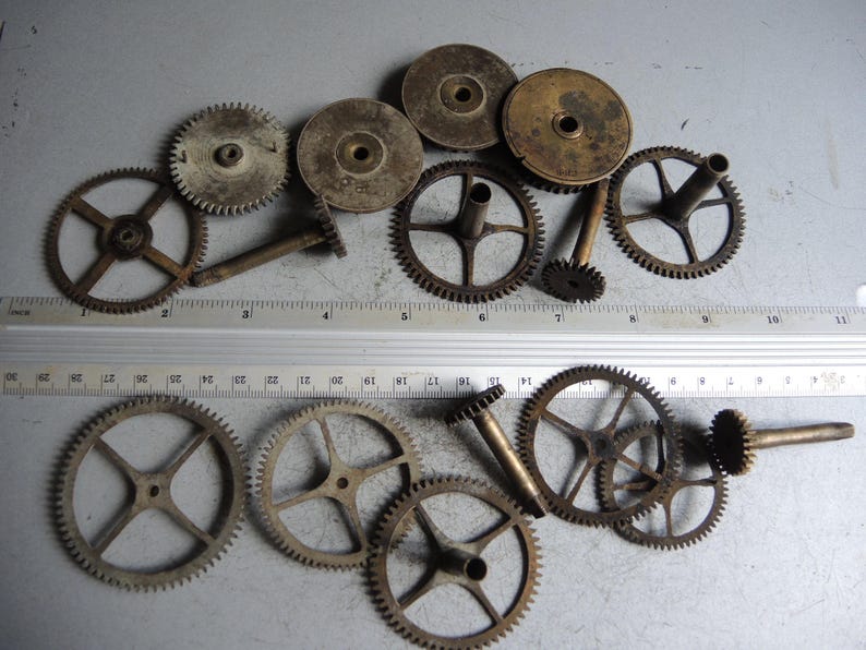 May include: A collection of vintage clock gears and components, showcasing various sizes and designs. The gears are made of metal and display a weathered, aged appearance. A ruler is placed near the gears for scale, with measurements in inches.