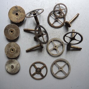 May include: A collection of vintage clock parts, including various sizes of brass gears and circular components. The gears have intricate teeth and are arranged on a gray surface, showcasing their aged appearance and mechanical detail.