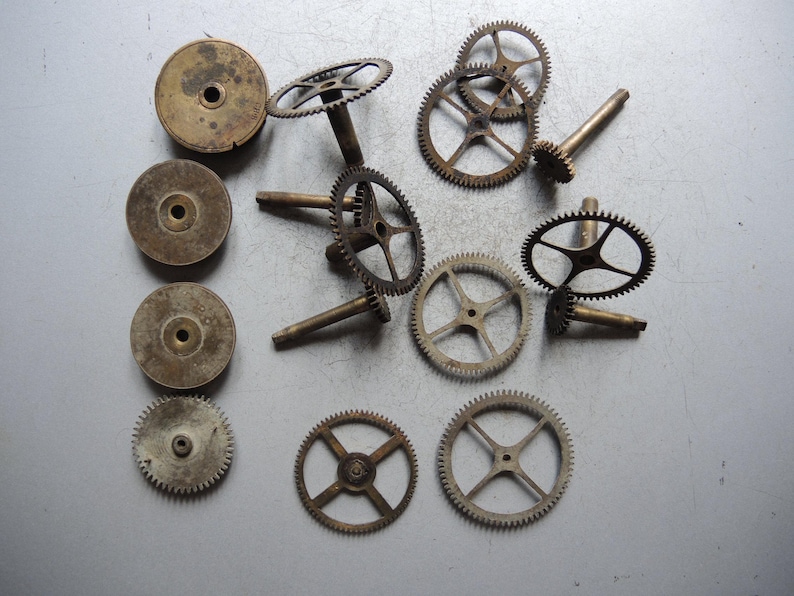 May include: A collection of vintage clock parts, including various sizes of brass gears and circular components. The gears have intricate teeth and are arranged on a gray surface, showcasing their aged appearance and mechanical detail.