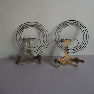 May include: Two antique clock balance wheels. Each has a gold-colored metal base with a decorative design and a black spiral spring. The clock parts are displayed against a neutral background.