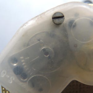 Op de afbeelding: Close-up van een doorzichtig plastic onderdeel met zichtbare interne tandwielen en een kleine schroef. Het onderdeel heeft een gebogen vorm en de tekst "S-0" is zichtbaar. De achtergrond is lichtblauw.