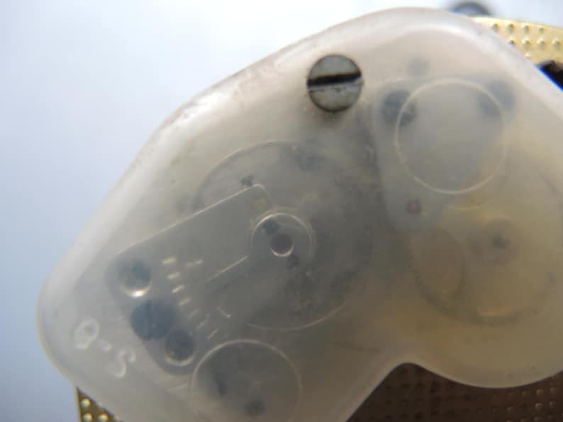 Op de afbeelding: Close-up van een doorzichtig plastic onderdeel met zichtbare interne tandwielen en een kleine schroef. Het onderdeel heeft een gebogen vorm en de tekst "S-0" is zichtbaar. De achtergrond is lichtblauw.