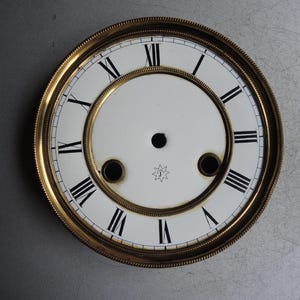 Könnte beinhalten: Ein Vintage-Zifferblatt mit einem goldfarbenen Metallrahmen und einem weißen Zifferblatt. Schwarze römische Ziffern markieren die Stunden. Das Zifferblatt hat ein zentrales Loch und zwei kleinere Löcher, mit einem sternförmigen Design unten.