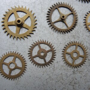 May include: Several small, brass-coloured gears with pointed teeth are scattered on a textured, light grey surface. The gears have different spoke designs and central holes. These components are likely for a clock or watch mechanism.
