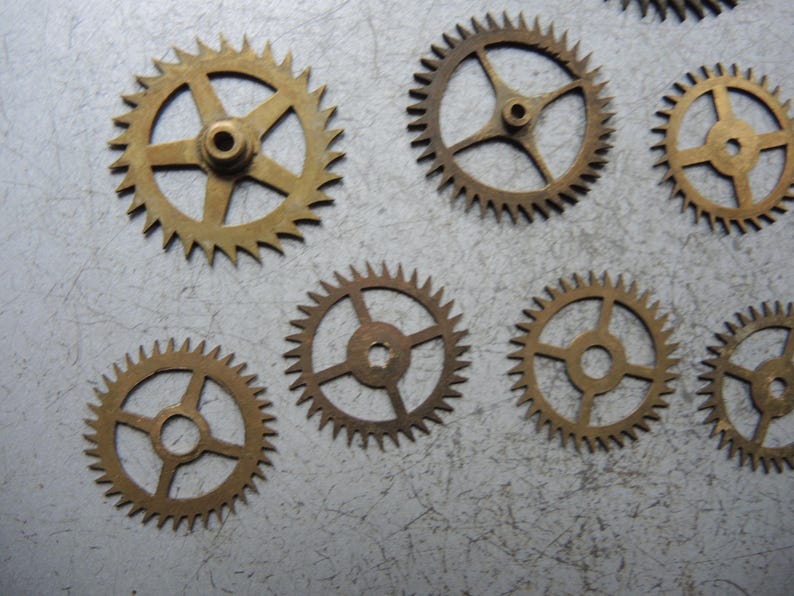 May include: Several small, brass-coloured gears with pointed teeth are scattered on a textured, light grey surface. The gears have different spoke designs and central holes. These components are likely for a clock or watch mechanism.