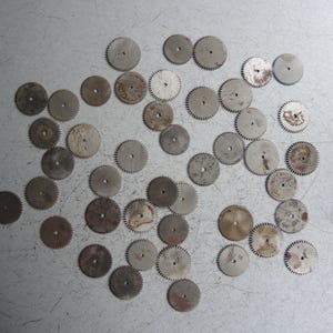 May include: A collection of small, circular metal gears with toothed edges, scattered on a light gray surface. The gears vary in size and show signs of wear, with some having a slightly rusty appearance. These components are likely from a watch or clock.