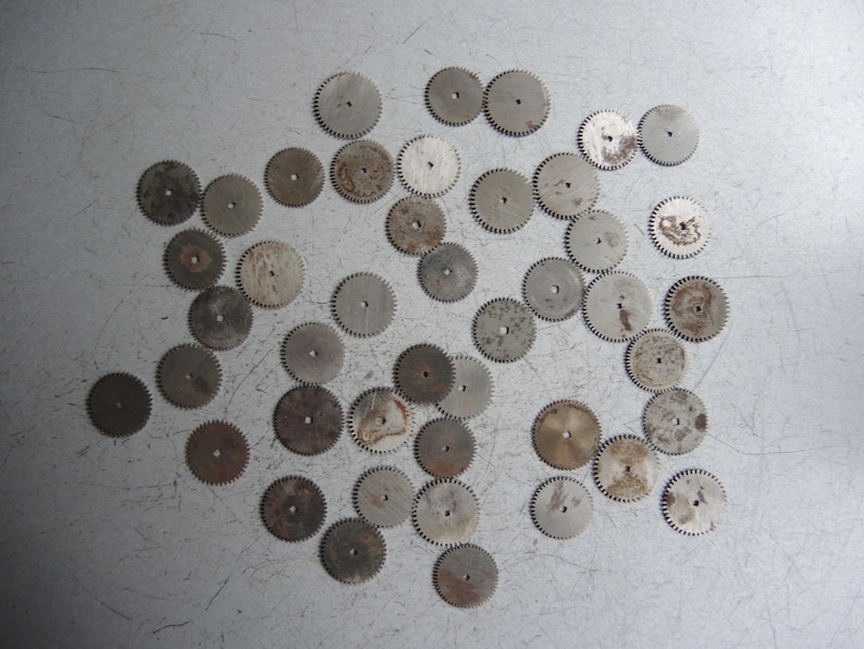 May include: A collection of small, circular metal gears with toothed edges, scattered on a light gray surface. The gears vary in size and show signs of wear, with some having a slightly rusty appearance. These components are likely from a watch or clock.