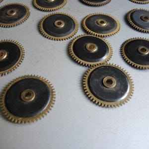 May include: A collection of small, circular watch gears. Each gear features a black center, a brass-colored outer ring with teeth, and a central brass-colored hole. The gears are scattered across a light-colored surface.