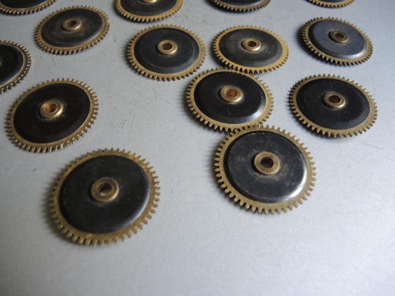 May include: A collection of small, circular watch gears. Each gear features a black center, a brass-colored outer ring with teeth, and a central brass-colored hole. The gears are scattered across a light-colored surface.