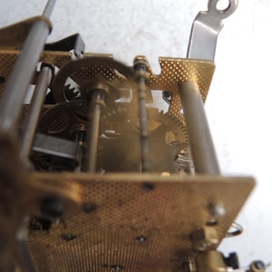 May include: Close-up of a clock mechanism with brass gears, plates, and metal rods. The gears are meshed together, and the plates have a textured, perforated design. The image is focused on the internal workings of the clock.