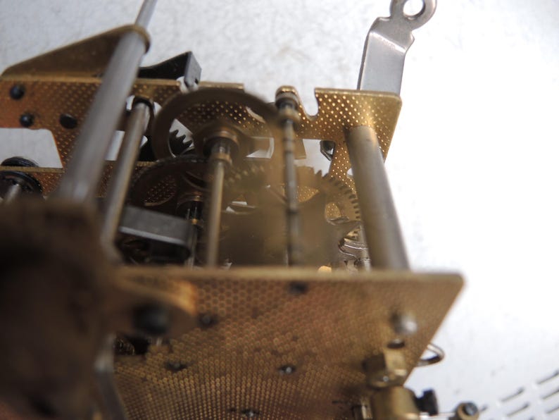 May include: Close-up of a clock mechanism with brass gears, plates, and metal rods. The gears are meshed together, and the plates have a textured, perforated design. The image is focused on the internal workings of the clock.