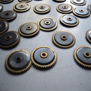 May include: A collection of small, circular watch gears. Each gear features a dark gray center, a brass-colored outer ring with teeth, and a central brass-colored hole. The gears are scattered across a light gray surface.