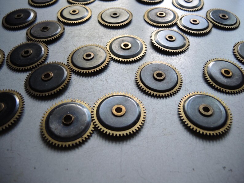 May include: A collection of small, circular watch gears. Each gear features a dark gray center, a brass-colored outer ring with teeth, and a central brass-colored hole. The gears are scattered across a light gray surface.