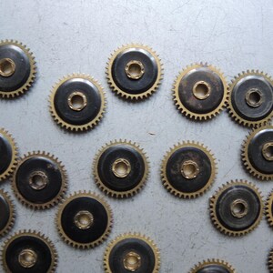 May include: A collection of small, circular watch gears with a dark center and gold-colored teeth. The gears are scattered across a light gray surface, showcasing their intricate design and mechanical detail.