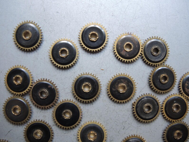 May include: A collection of small, circular watch gears with a dark center and gold-colored teeth. The gears are scattered across a light gray surface, showcasing their intricate design and mechanical detail.