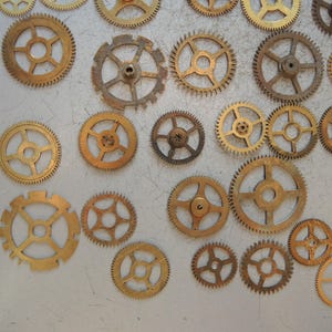 以下が含まれることがあります： さまざまなサイズの真鍮製歯車のコレクション。 金色の歯車は、明るい灰色の表面に配置されています。 いくつかの歯車は、ヴィンテージ感を高める摩耗の兆候を示しています。