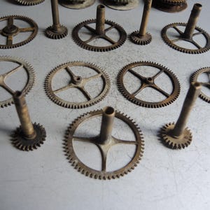 May include: A collection of antique clock gears in various sizes and designs. The gears are made of metal with a tarnished, aged appearance. Some gears have spokes, while others have solid centers. The gears are arranged on a light gray surface.
