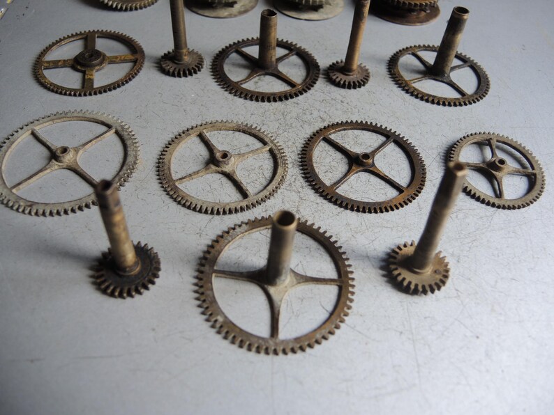 May include: A collection of antique clock gears in various sizes and designs. The gears are made of metal with a tarnished, aged appearance. Some gears have spokes, while others have solid centers. The gears are arranged on a light gray surface.