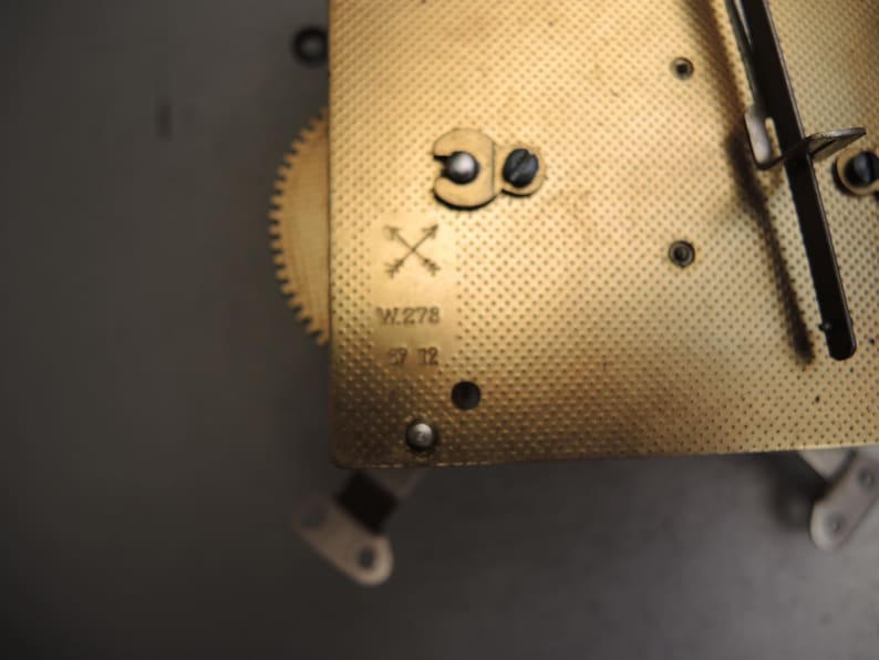 May include: Close-up of a vintage clock mechanism with a gold-colored, textured plate. The plate has a series of small holes and is marked with the text "W.278" and "29 12". A small gear and other mechanical components are visible.