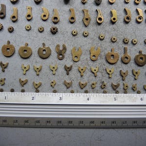 May include: A collection of small, antique brass clock parts, including gears, levers, and shaped components. A metal ruler with inch markings is placed below the parts for scale. The parts are arranged on a gray surface.