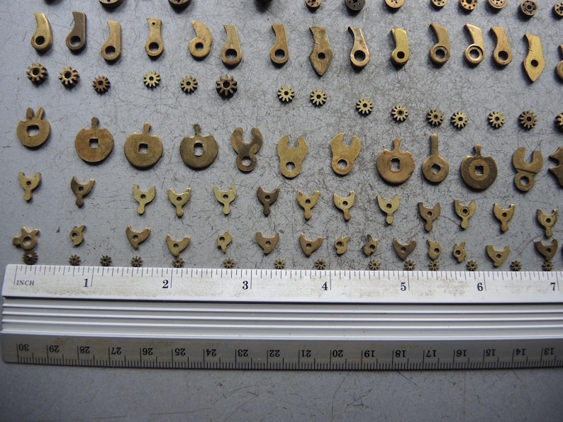 May include: A collection of small, antique brass clock parts, including gears, levers, and shaped components. A metal ruler with inch markings is placed below the parts for scale. The parts are arranged on a gray surface.