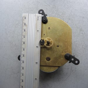 Op de afbeelding: Uurwerk met messing tandwielen en een metalen liniaal met metingen tot 76 cm. Het uurwerk heeft een goudkleurige plaat en zwarte montagebeugels. De liniaal is zilverkleurig met zwarte markeringen.