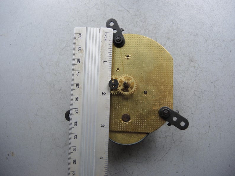 Op de afbeelding: Uurwerk met messing tandwielen en een metalen liniaal met metingen tot 76 cm. Het uurwerk heeft een goudkleurige plaat en zwarte montagebeugels. De liniaal is zilverkleurig met zwarte markeringen.