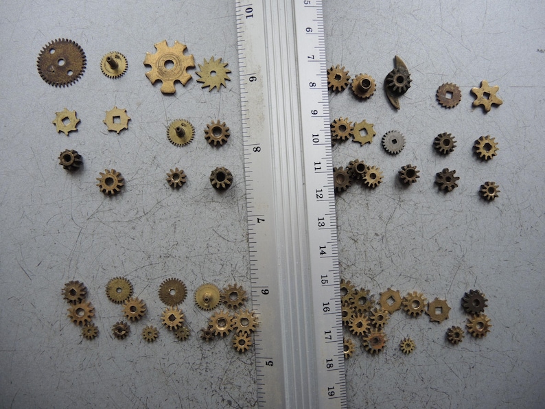 May include: A selection of small, brass-coloured gears in diverse shapes and sizes, including circular, star-shaped, and square-holed designs. A ruler with centimetre markings is placed next to the gears. The gears are scattered on a grey surface.