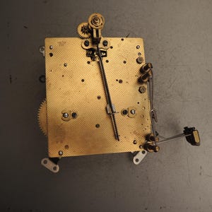 May include: A vintage clock mechanism featuring brass gears and a gold-coloured metal base. The movement includes a pendulum rod, springs, and various components. The clock's intricate design is visible against a grey background.