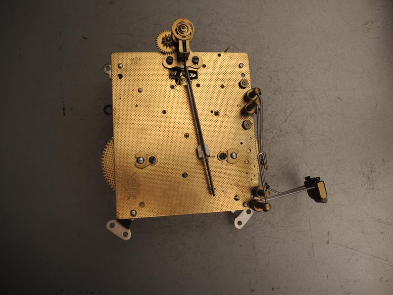 May include: A vintage clock movement with brass gears and a gold-toned metal base. The mechanism includes a pendulum rod, springs, and various components. The clock's intricate design is visible against a gray background.