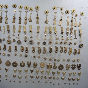 May include: A collection of small, vintage brass watch parts, including gears, levers, and other intricate components. The parts are arranged on a gray surface, showcasing their detailed designs and aged patina. A variety of shapes and sizes are present.