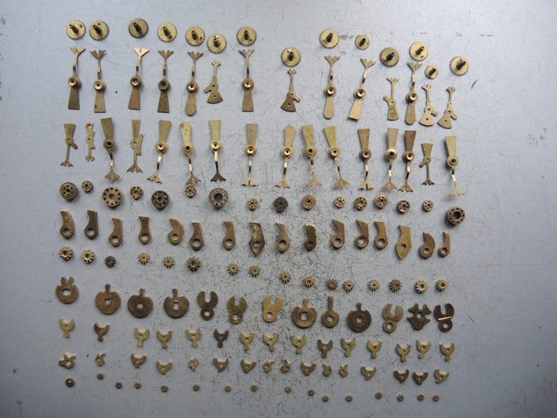 May include: A collection of small, vintage brass watch parts, including gears, levers, and other intricate components. The parts are arranged on a gray surface, showcasing their detailed designs and aged patina. A variety of shapes and sizes are present.