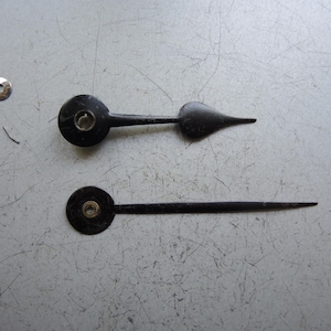 Op de afbeelding: Twee zwarte wijzers en een kleine metalen ring. Eén wijzer heeft een puntig uiteinde, de andere is recht. De wijzers liggen op een lichtgrijze ondergrond.