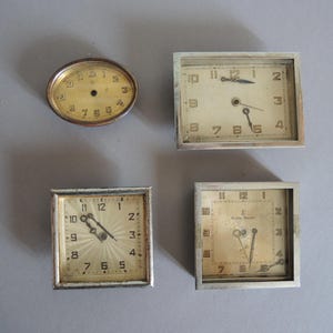 May include: Four vintage clocks with silver frames and cream-colored faces are displayed. One is oval, while the others are square or rectangular. The clock faces feature black numerals and hands, showcasing a classic design.