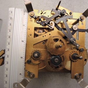 May include: Close-up of a clock mechanism with brass gears, levers, and a metal ruler. The ruler measures up to 30 inches. The clock's intricate design features various gears and components, showcasing its internal workings.
