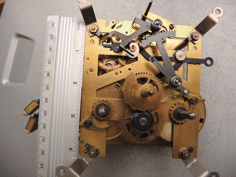 May include: Close-up of a clock mechanism with brass gears, levers, and a metal ruler. The ruler measures up to 30 inches. The clock's intricate design features various gears and components, showcasing its internal workings.