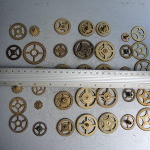 May include: A collection of various sized brass gears, some with spokes, arranged on a gray surface. A metal ruler, marked in inches, is placed across the gears. The gears range in size, with the largest being approximately 2 inches in diameter.
