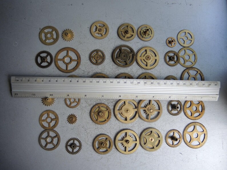 May include: A collection of various sized brass gears, some with spokes, arranged on a gray surface. A metal ruler, marked in inches, is placed across the gears. The gears range in size, with the largest being approximately 2 inches in diameter.