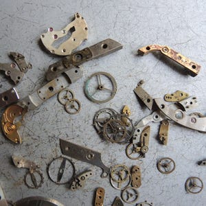 K&ouml;nnte beinhalten: Eine Sammlung zerlegter Uhrenteile, darunter Zahnr&auml;der, Hebel und Platten, auf einer grauen Oberfl&auml;che verstreut. Die Komponenten bestehen aus Metall mit verschiedenen Silber-, Gold- und Bronzet&ouml;nen. Das Bild zeigt die komplizierten Details von Uhrenmechanismen.