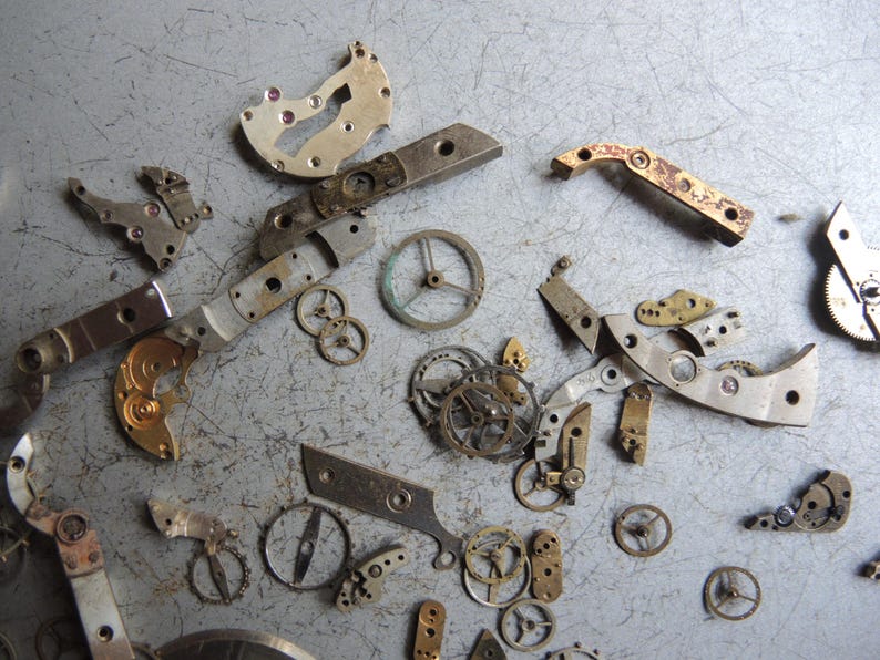 K&ouml;nnte beinhalten: Eine Sammlung zerlegter Uhrenteile, darunter Zahnr&auml;der, Hebel und Platten, auf einer grauen Oberfl&auml;che verstreut. Die Komponenten bestehen aus Metall mit verschiedenen Silber-, Gold- und Bronzet&ouml;nen. Das Bild zeigt die komplizierten Details von Uhrenmechanismen.