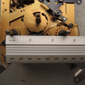 May include: Close-up of a clock mechanism with brass gears and a silver ruler. The ruler measures up to 4 inches. The gears are intricately designed, showing the inner workings of a clock. The mechanism is set against a gray background.