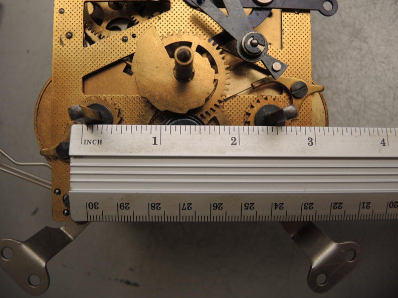 May include: Close-up of a clock mechanism with brass gears and a silver ruler. The ruler measures up to 4 inches. The gears are intricately designed, showing the inner workings of a clock. The mechanism is set against a gray background.