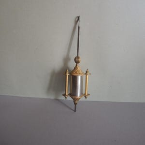 May include: A vertical clock pendulum featuring a central cylindrical silver-toned weight flanked by two thin gold-toned rods. The assembly is topped and bottomed with ornate gold-toned decorative finials and a slender metal suspension rod.