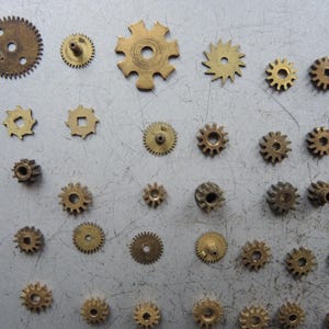 May include: A selection of small brass gears in various sizes and designs. The gears have different numbers of teeth and central holes. Some are circular, others more complex. The gears are scattered on a grey surface. Measurements are in centimetres.